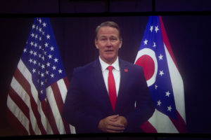Picture of man speaking in front of the US and Ohio flag.